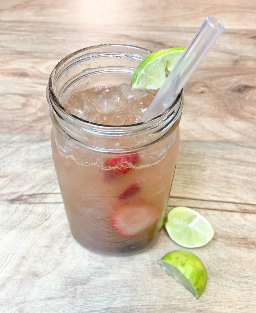 A mason jar filled with ice, strawberries, cherries and a drink with slices of lime
