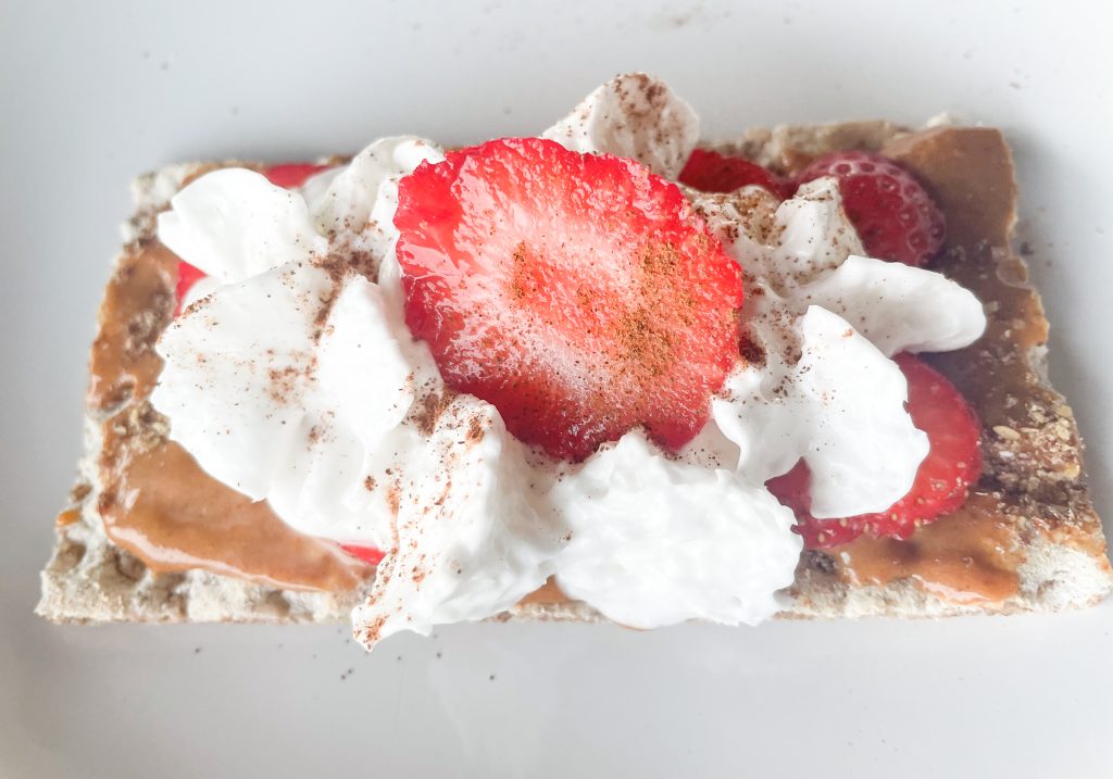 a wasa cracker on a white surface topped with coach kanada peanut butter, strawberries, cinnamon, and almond whipped cream.